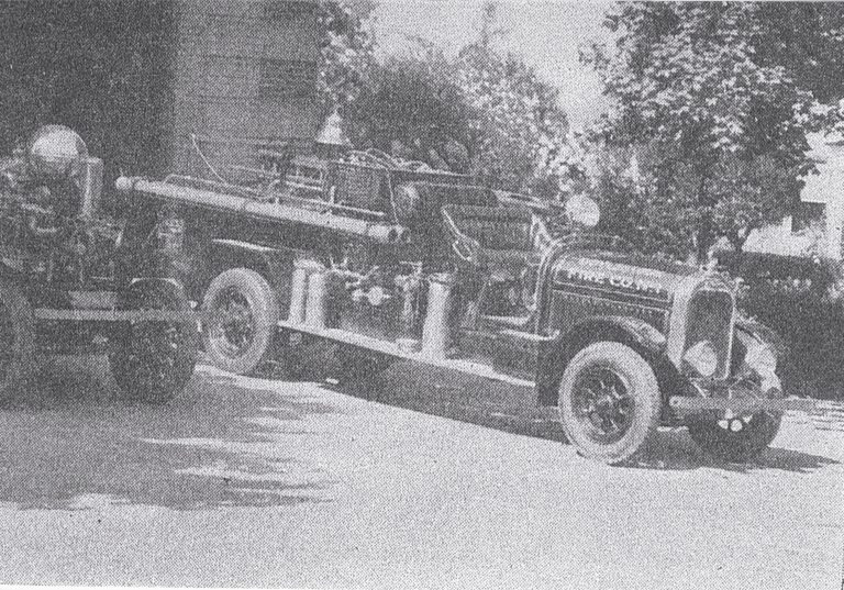 Black and white photo of a vintage fire truck parked outdoors, surrounded by trees and a building in the background; the truck has ladders and equipment on its sides.