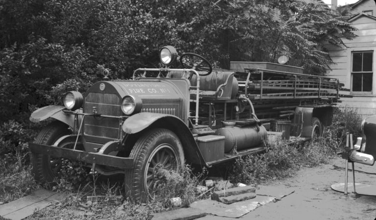 A vintage fire truck with "Fire Co. No. 1" on the front sits overgrown with weeds and plants, surrounded by old tools and an abandoned chair, beside a small building. The image is in black and white.