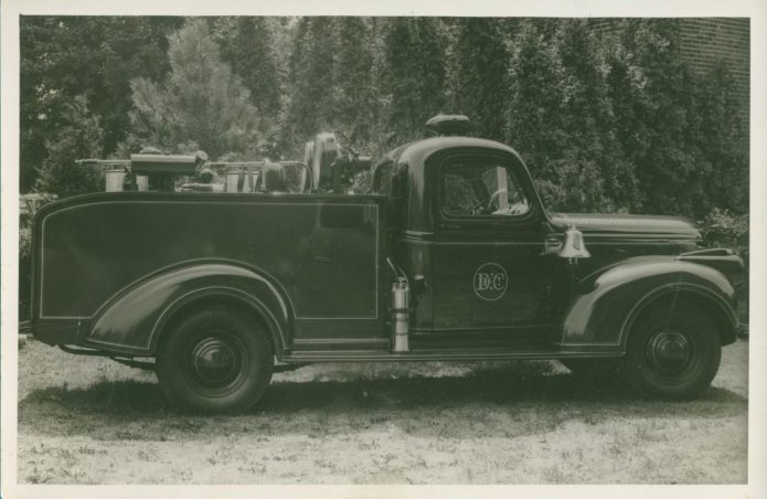 Black-and-white photo of a vintage fire truck parked on grass, with equipment visible in the truck bed and the letters "DPC" on the door. Shrubs and trees are in the background.