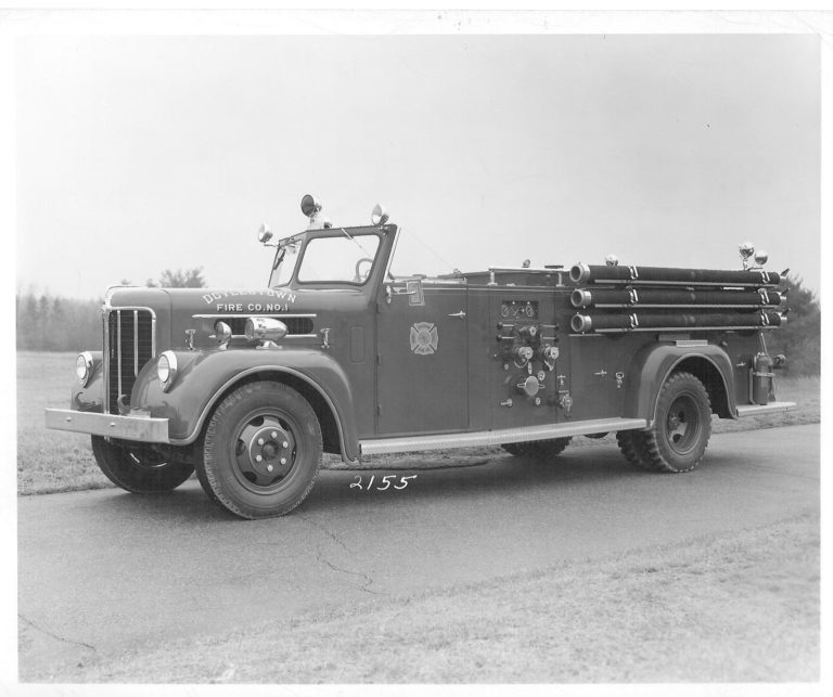 Black-and-white photo of a vintage fire truck parked on a paved road, equipped with hoses along the side and emergency lights on top. "Fire Co. No. 1" is visible on the side. The background shows grass and trees.