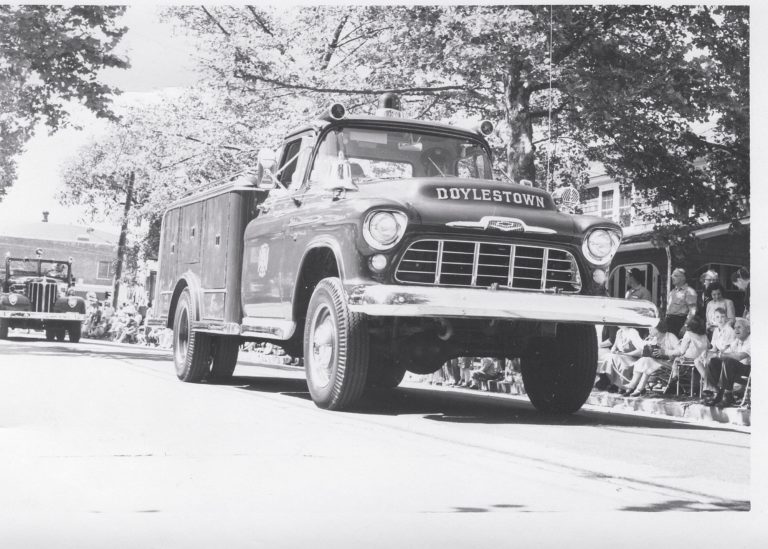 A vintage fire truck labeled “Doylestown” drives down a street in a parade, with people sitting on the curb watching and trees lining the background. The image is black and white.