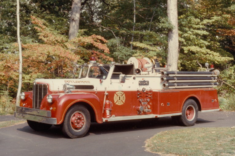 A vintage red and white fire truck with hoses and equipment is parked on a paved road, surrounded by green trees and foliage in the background.