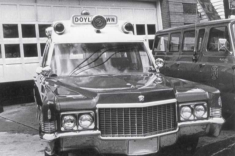 A vintage black-and-white photo of an old Cadillac ambulance with “DOYLESTOWN” on a sign above the windshield, parked in front of a garage next to a van.