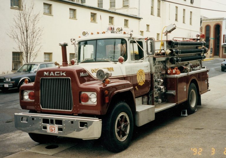 A vintage red and white MACK fire truck is parked on a city street near a beige building. Fire hoses and firefighting equipment are visible on the truck.