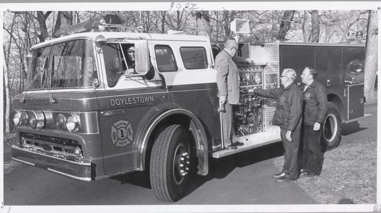 Black and white photo of a Doylestown fire truck with three men standing beside it and one man sitting in the driver’s seat. The scene takes place outdoors with trees in the background.