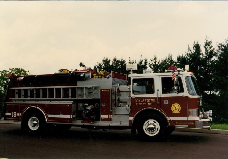 A red and white fire truck labeled "Doylestown Fire Co No 1" is parked on a road with trees and a cloudy sky in the background. The truck has various equipment and an American flag attached to the cab.