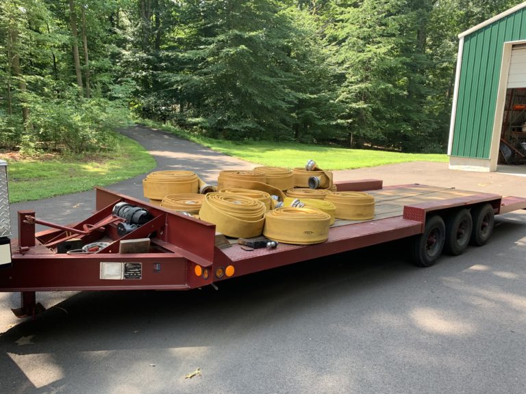 A flatbed trailer with three axles is parked on a driveway, carrying several coiled yellow fire hoses and some metal equipment. The scene is outdoors near a green building and surrounded by trees.