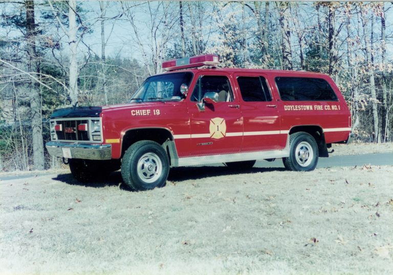 A red Doylestown Fire Co. No. 1 SUV marked "Chief 19" is parked on grass near trees, with emergency lights on top and a fire department emblem on the door.