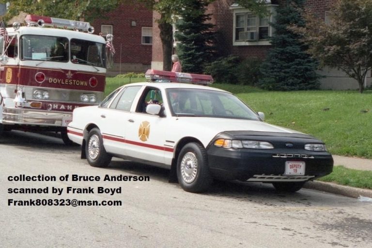 A white sedan with emergency lights and a fire department emblem is parked in front of a fire truck labeled "Doylestown" on a residential street with grass and brick buildings in the background.