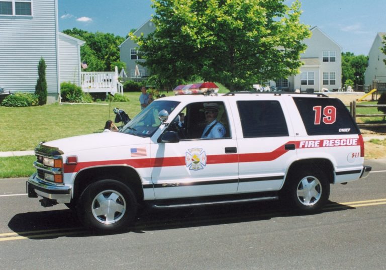 A white SUV marked "FIRE RESCUE" with the number 19 and emergency lights on the roof is parked on a suburban street. A uniformed person is visible inside, and houses and trees are in the background.