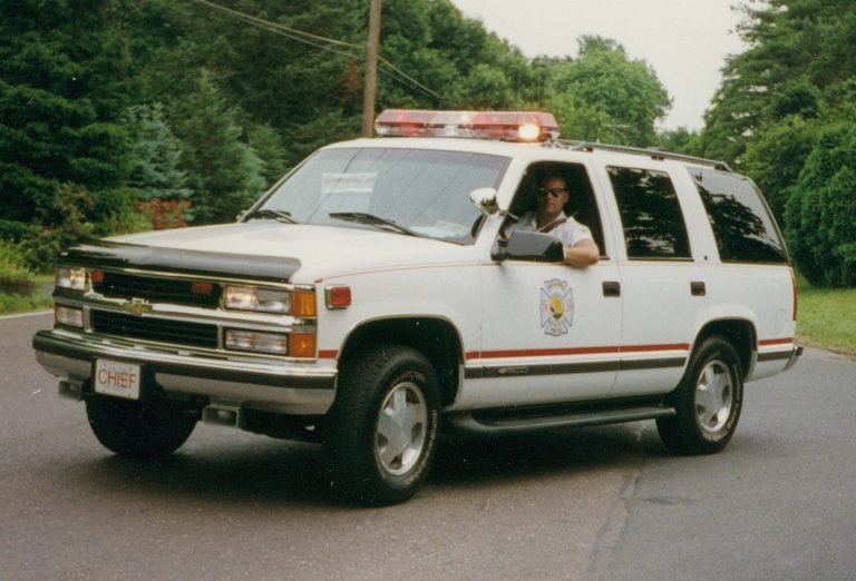 A white Chevrolet SUV with emergency lights and a "CHIEF" license plate is parked on a suburban street. A person wearing sunglasses is sitting in the driver's seat. Trees and greenery are in the background.