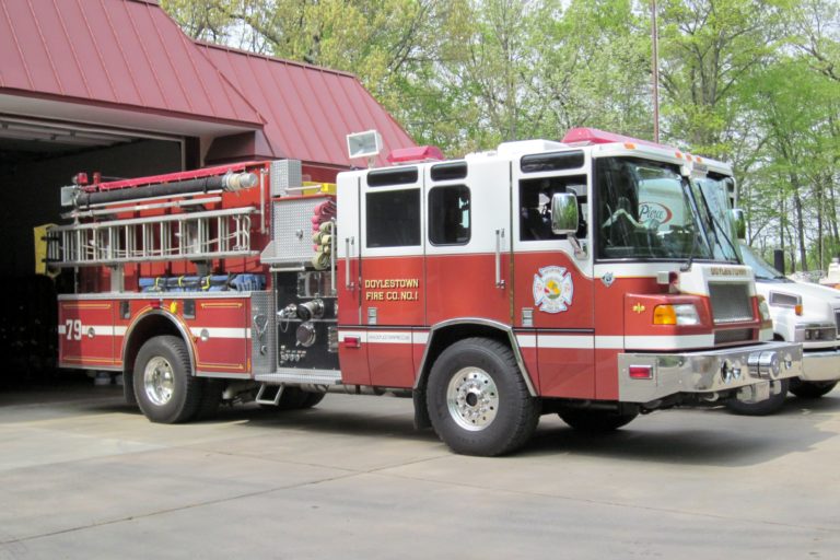 A red and white fire truck from Doylestown Fire Company is parked outside a building with a red roof on a sunny day. Trees with green leaves are visible in the background.