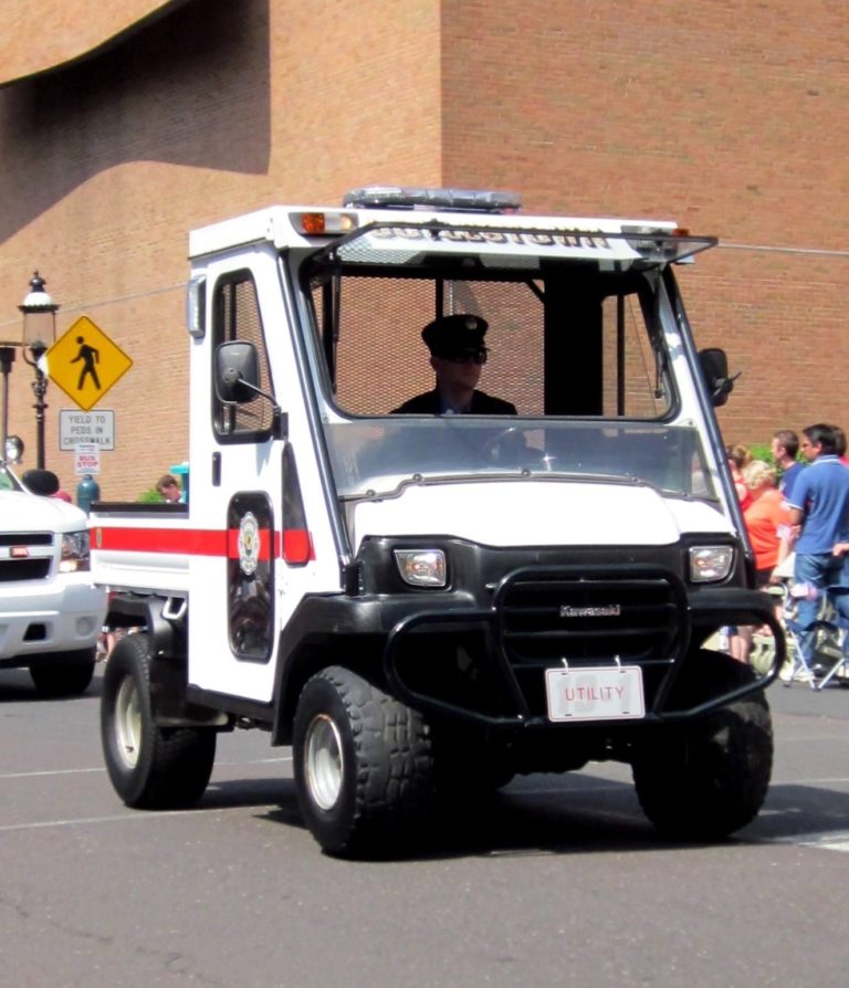 A police officer drives a small utility vehicle with flashing lights during a parade, with a crowd and brick buildings visible in the background.