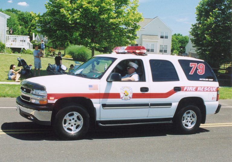 A white SUV marked "Fire Rescue" with the number 79 and emergency lights on the roof drives down a suburban street during the day. People sit and stand on the grassy roadside in the background.