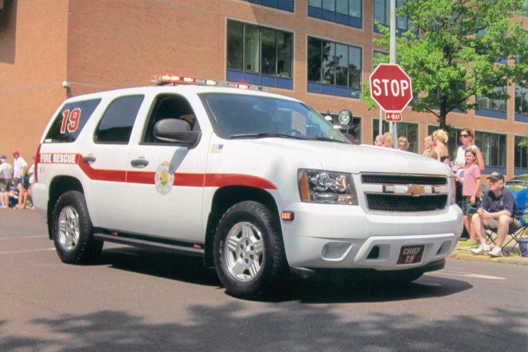 A white fire rescue SUV with emergency lights and the number 19 on the side drives in a parade. People stand and sit on the sidewalk; a stop sign and a brick building are visible in the background.