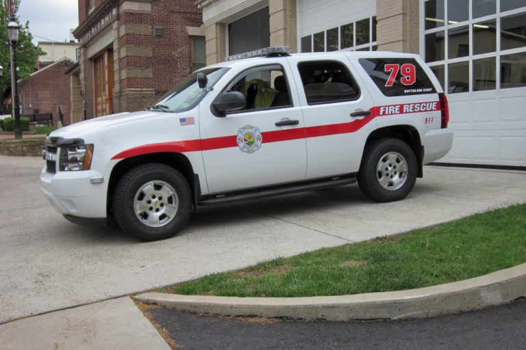 A white fire rescue SUV with red stripes and the number 79 is parked outside a fire station. The vehicle has emergency lights on the roof and fire department insignia on the front door.