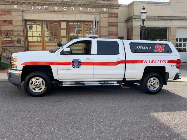 A white fire rescue utility truck labeled "UTILITY 19" and "FIRE RESCUE" is parked in front of a fire station building with large windows and an arched doorway.