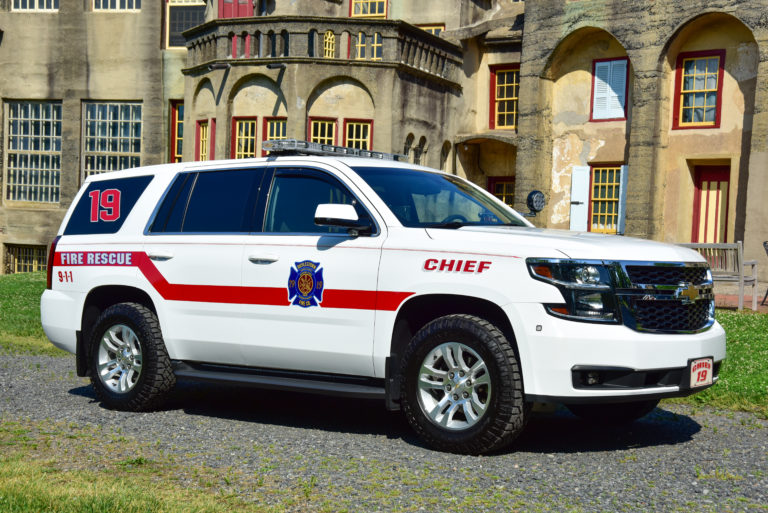A white fire rescue SUV marked “CHIEF” and “19” is parked on grass in front of a historic stone building with arched windows and red trim. The vehicle has red stripes and emergency insignias on the side.