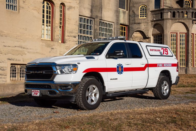 A white RAM pickup truck marked "Battalion 79 Fire Rescue" with red stripes and a fire department emblem is parked on gravel in front of a large stone building with many windows.