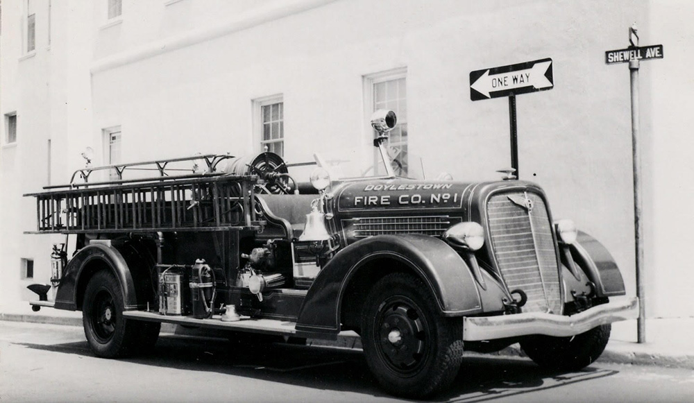 Black and white photo of a vintage Doylestown Fire Co. No. 1 fire truck parked on a street corner near a "One Way" sign and a street sign labeled "Shewell Ave.