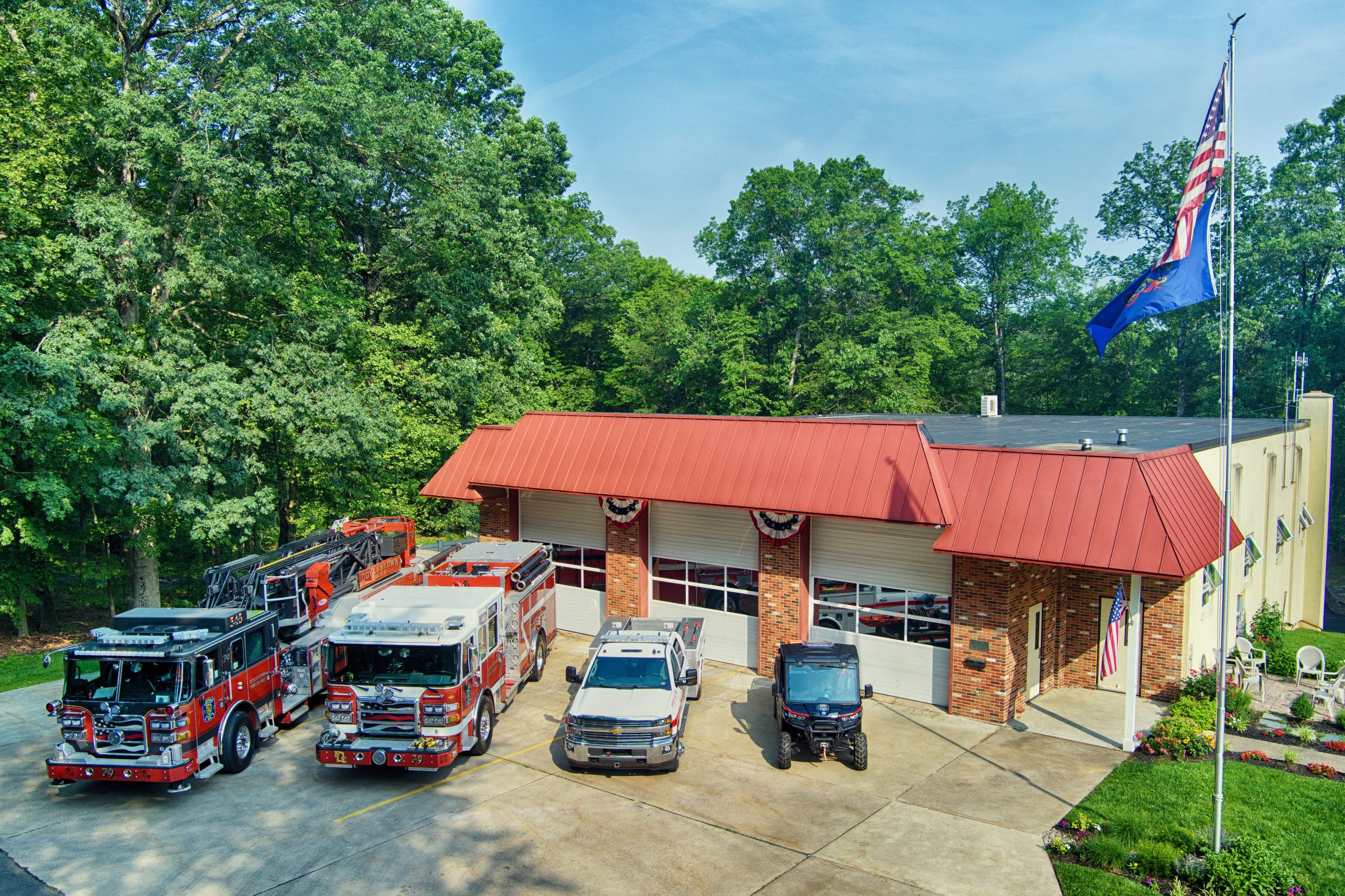 A fire station with a red roof and three garage bays stands surrounded by trees. Four emergency vehicles and two utility vehicles are parked in front, and a flagpole with American and blue flags is visible on a sunny day.