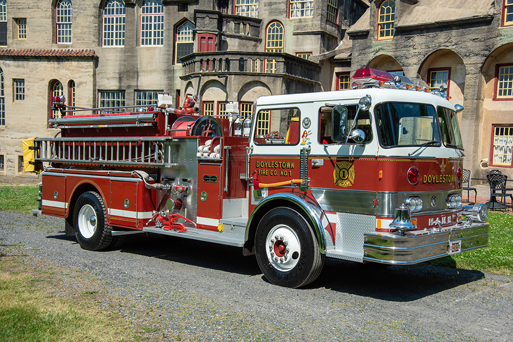 A vintage red and white fire truck from Doylestown Fire Co. No. 1 is parked on grass in front of a historic, multi-story building with arched windows and decorative stonework.