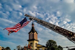 A large American flag hangs from a raised Doylestown Fire Co. No. 1 ladder truck, with a church steeple and trees visible below against a partly cloudy sky.