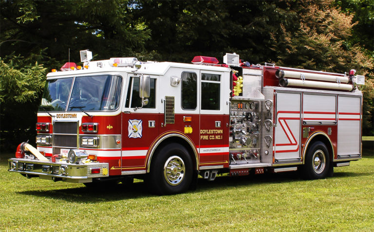 A red and white fire truck with "Doylestown Fire Co. No. 1" written on the side is parked on green grass, surrounded by trees on a sunny day.