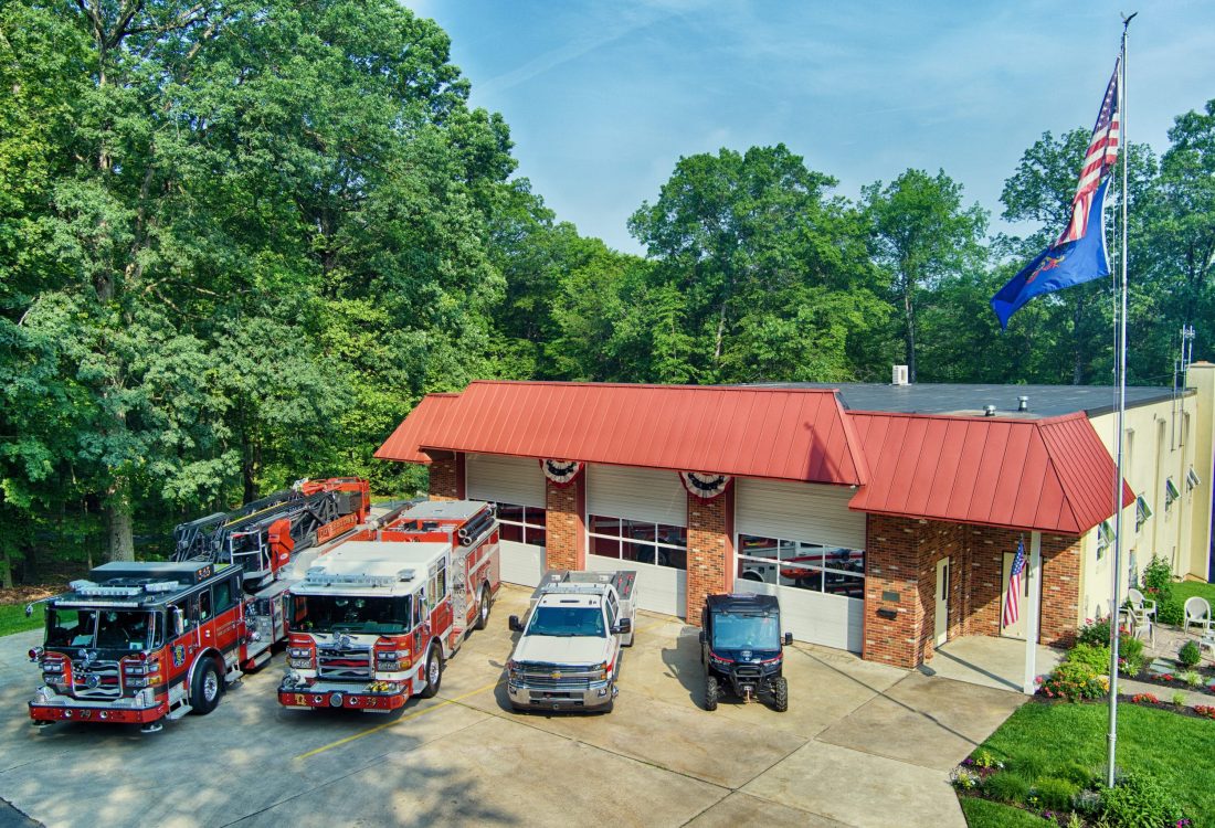 A fire station with a red roof and three garage bays stands surrounded by trees. Four emergency vehicles and two utility vehicles are parked in front, and a flagpole with American and blue flags is visible on a sunny day.